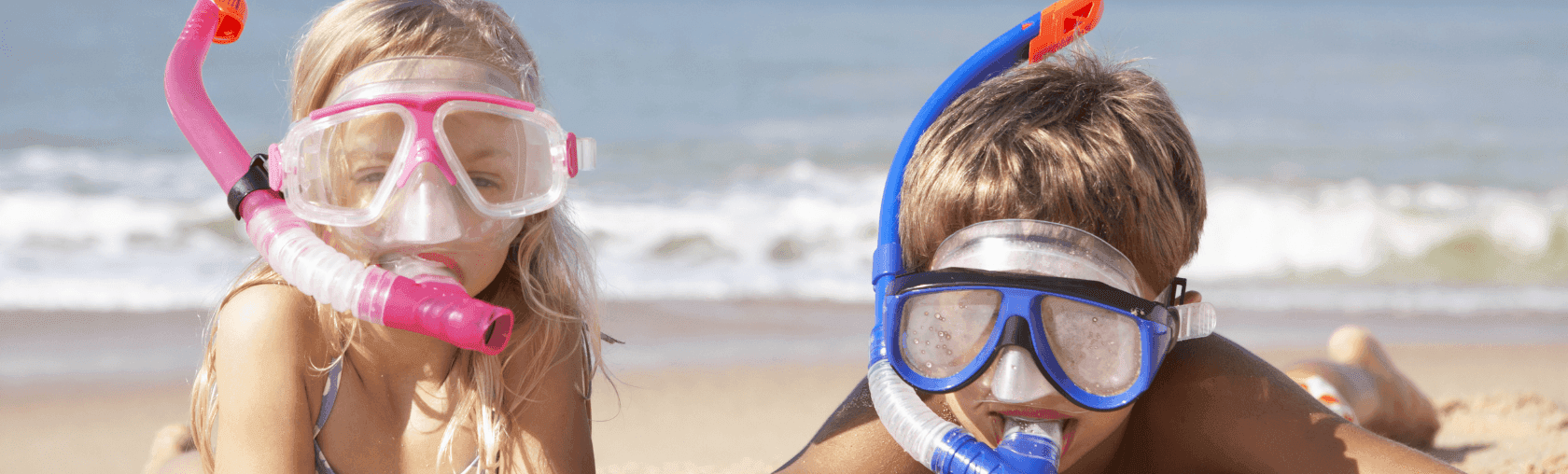 Two children wearing pink and blue snorkeling masks and snorkels lie on a sandy beach.
